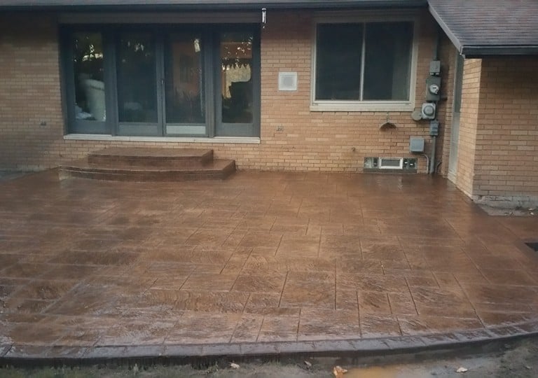 Custom brown stamped concrete patio with decorative borders and matching stairs behind a brick house.