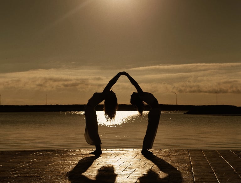 Silhouette of two people practicing yoga together on a coastal pier during a golden sunset.