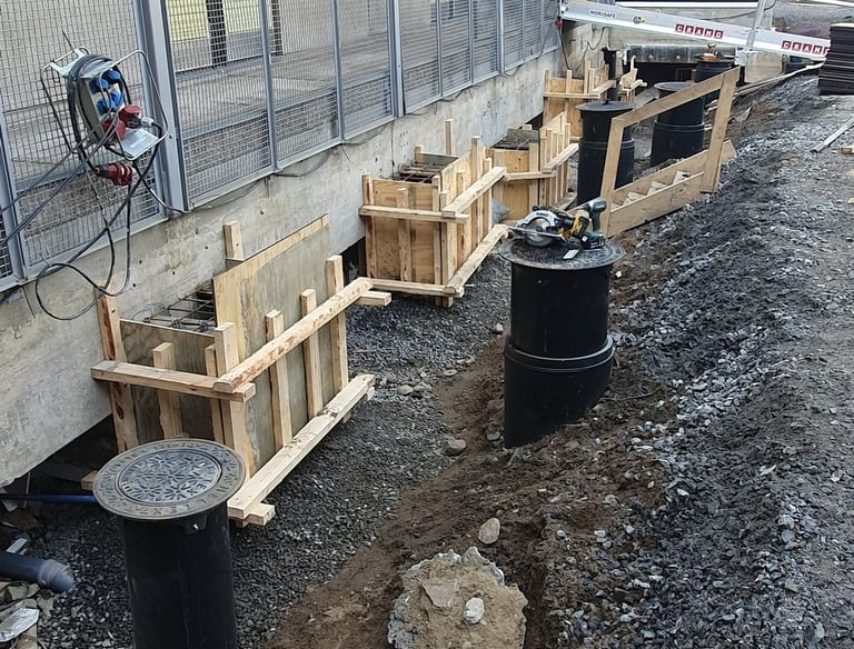 Foundation construction with wooden formwork and black drainage pipes next to a modern building.