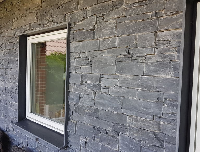Moderne ydervæg på hus med grå stablet stenbeklædning og hvidindrammede vinduer.