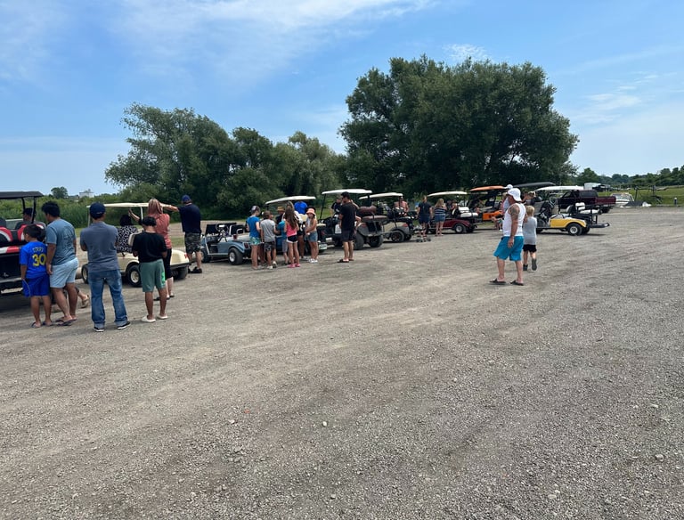A group of people and families gathering by a row of parked golf carts in a gravel parking lot outdoors.