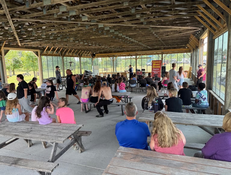 Families gather at a campground pavilion for a live magic show performance on a sunny afternoon.