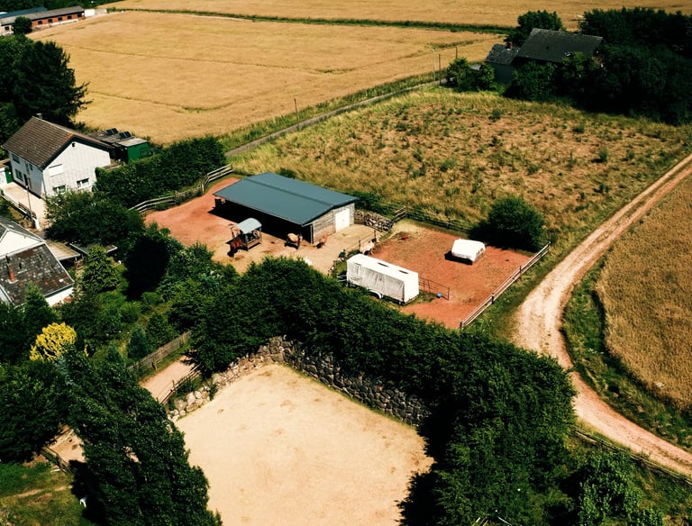 Luftaufnahme eines ländlichen Hofes mit Stall, umzäuntem Reitplatz und angrenzenden Feldern