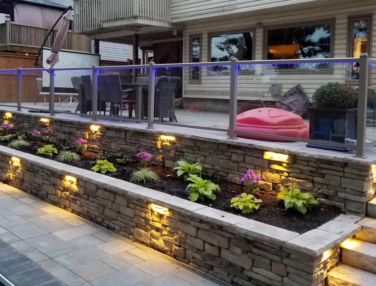 Illuminated stacked stone retaining wall with garden bed and glass railing on a backyard patio.