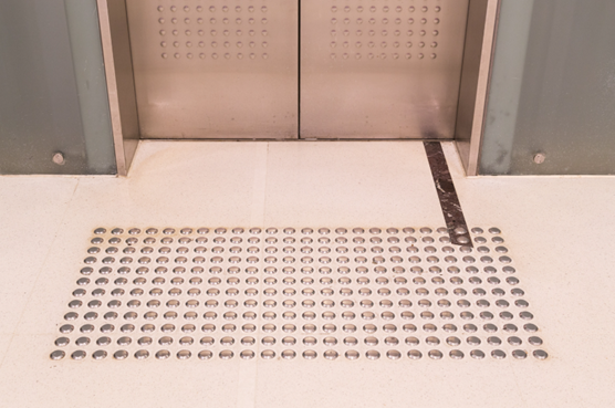 parte inferior de uma porta fechada de elevador e a sua frente no piso, piso tátil instalado e uma régua medindo espaço