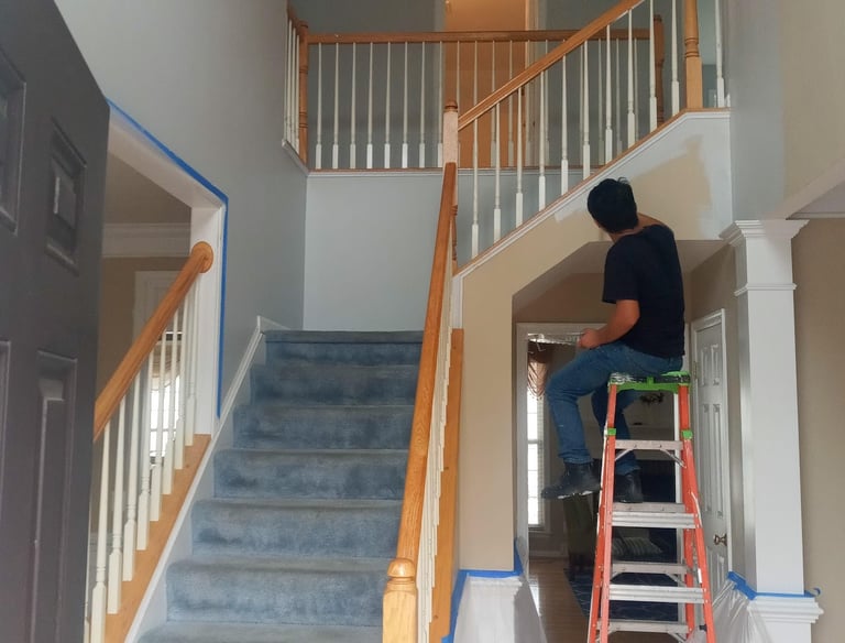 A person working on home renovation, standing on a ladder in a room with white walls and a staircase