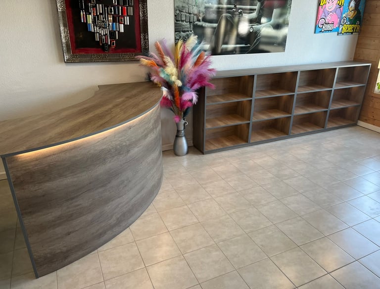 Modern curved wooden reception desk with matching shelving unit and colorful pampas grass decor.