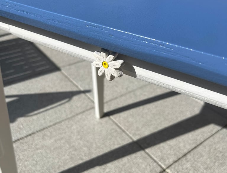 White daisy tablecloth weight clipped to a blue patio table on a sunny outdoor terrace.