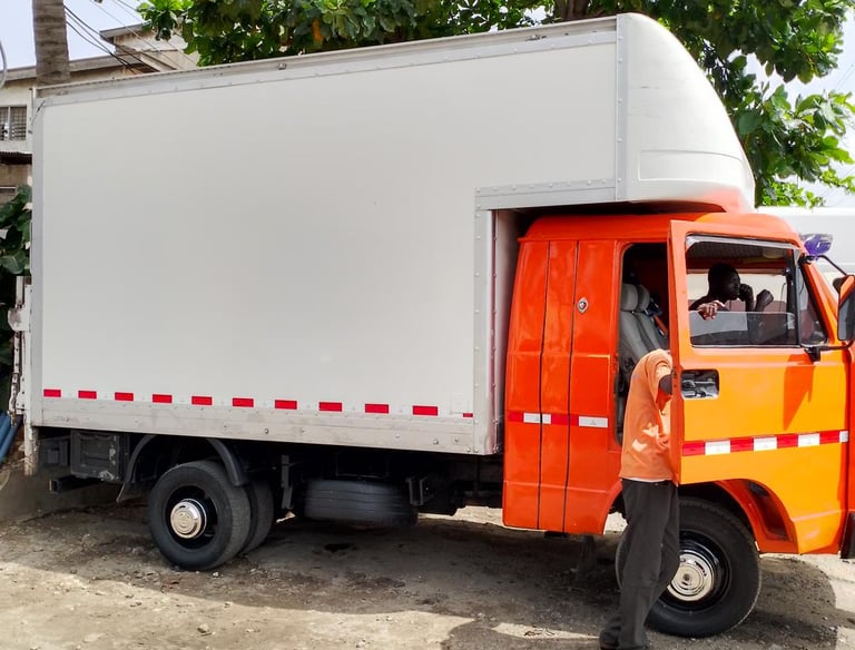 An orange cab delivery truck with a white enclosed cargo box and red reflective stripes, parked on a