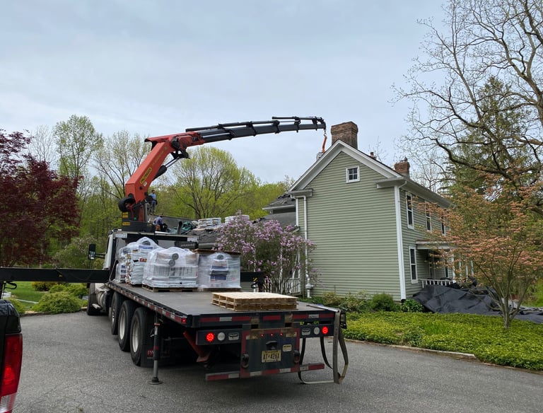 A knuckle boom crane truck delivering roofing materials to the roof of a green two-story residential house.