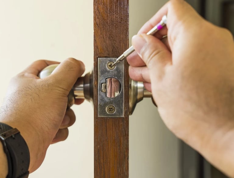 A person using a small screwdriver to repair a metal door latch on a wooden door frame.