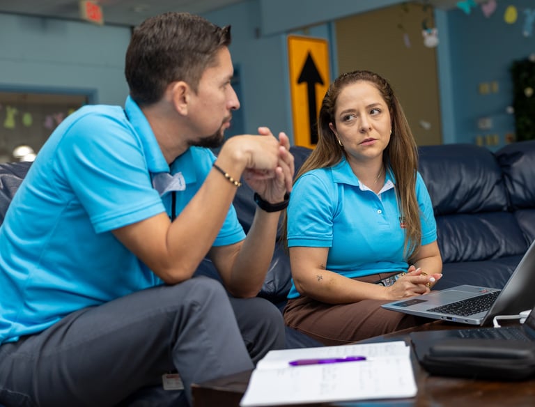 Two colleagues in blue polo shirts discuss project data over a laptop in a modern office lounge.