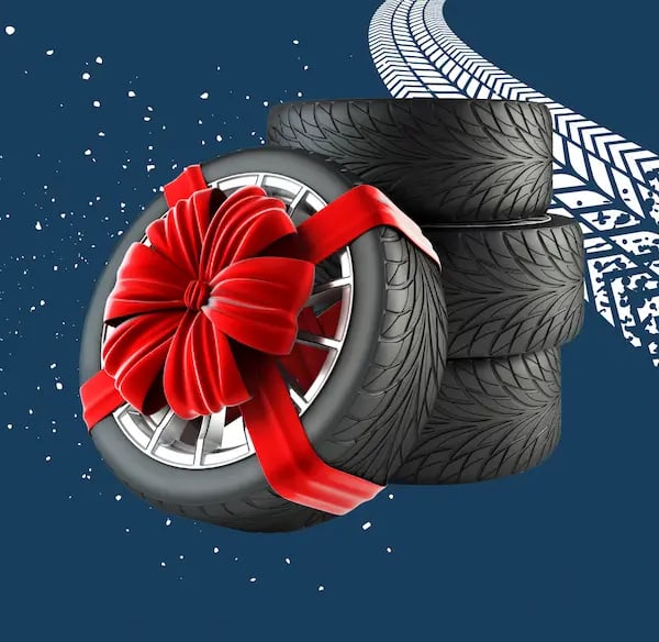 Cuatro neumáticos de coche apilados y un quinto neumático con una llanta plateada y un gran lazo de regalo rojo, todo sobre u