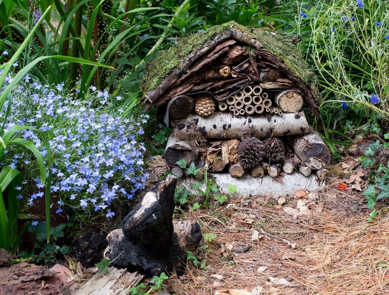 Handmade wooden insect hotel in a lush garden with blue wildflowers and a moss-covered roof.