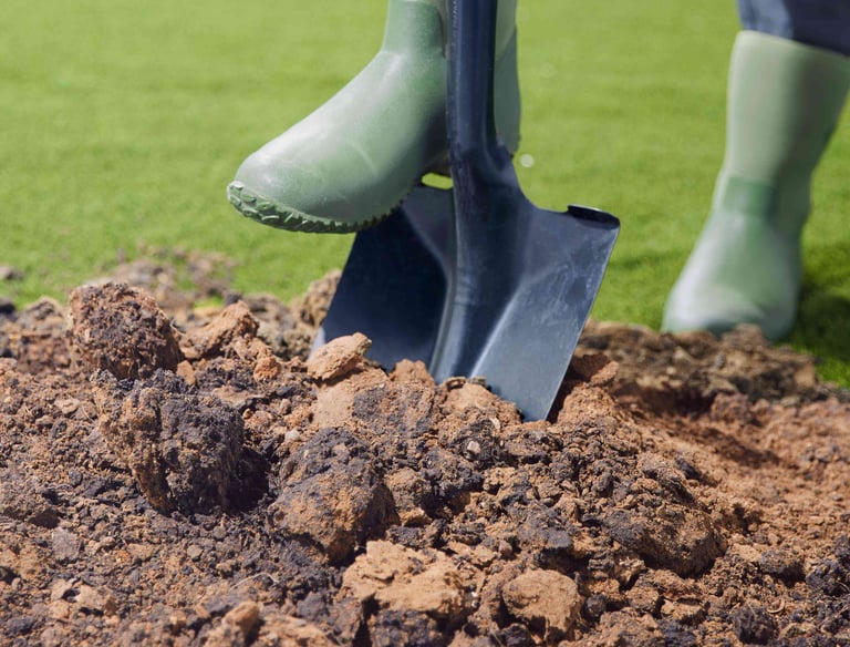 A gardener in green rubber boots uses a metal shovel to dig into a pile of garden soil on a lawn.