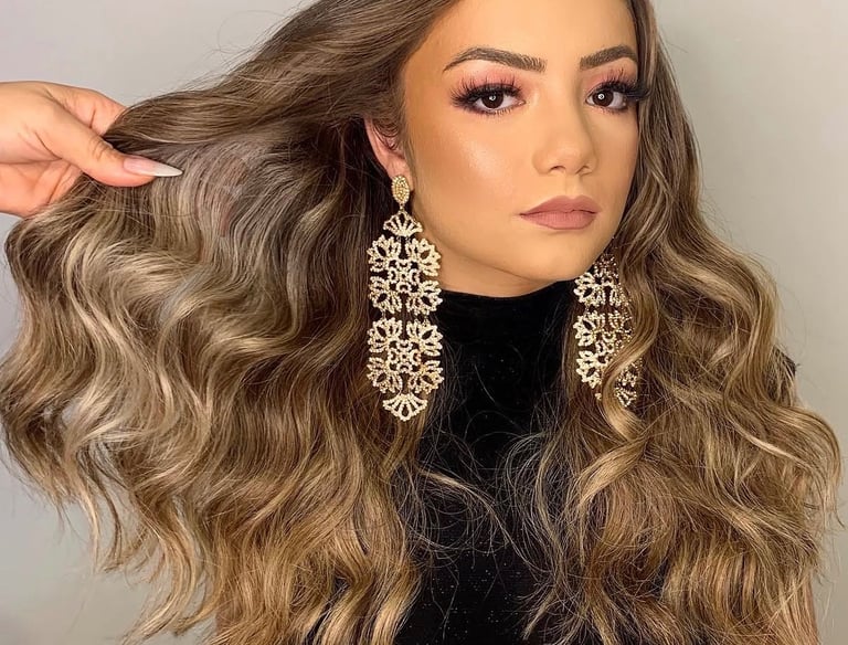 Woman with long wavy balayage hair, glamorous pink eyeshadow, and gold statement chandelier earrings.