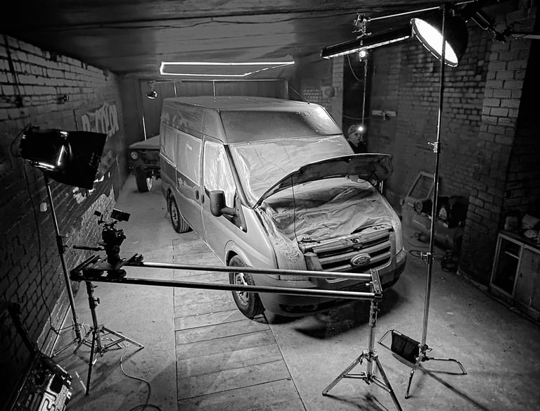 Filmavimo aikštelė garaže, kurioje yra furgonas su studijiniu apšvietimu ir kameros slideriu.
