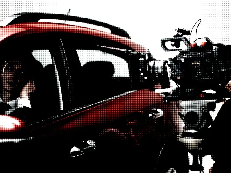 Film camera facing actor in car.