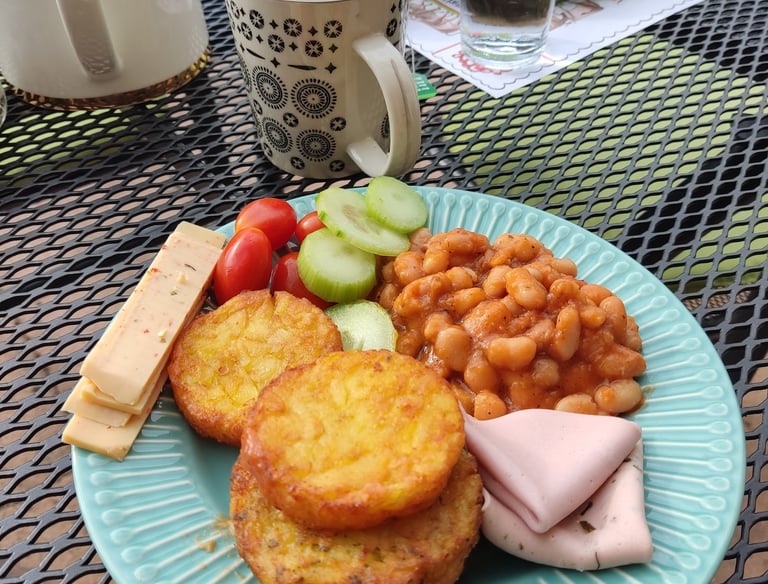 A hearty breakfast plate with hash browns, baked beans, ham, and fresh vegetables on an outdoor patio table.