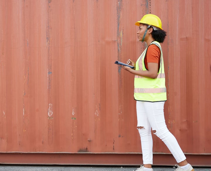 Female logistics manager in hard hat using a digital tablet by a red shipping container.