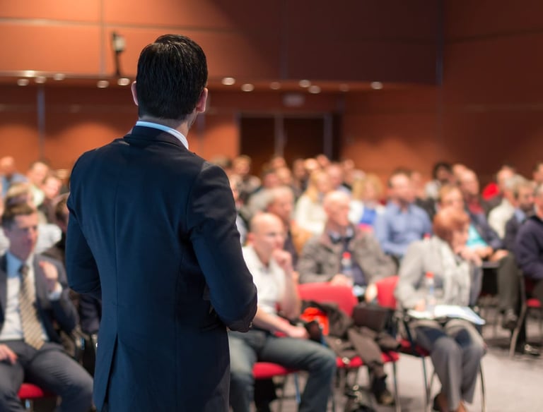 Professional public speaker in a business suit giving a presentation to a large audience at a conference.