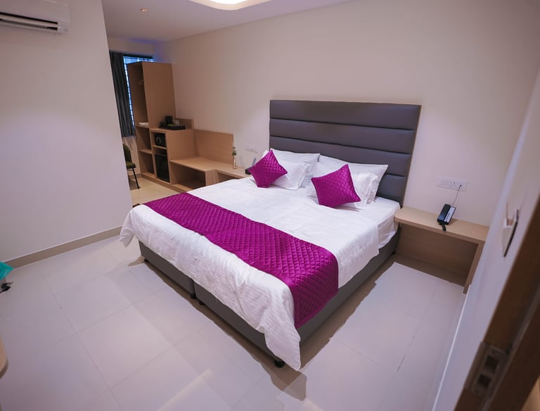 Modern hotel room with a queen bed, grey upholstered headboard, and purple decorative pillows and runner.