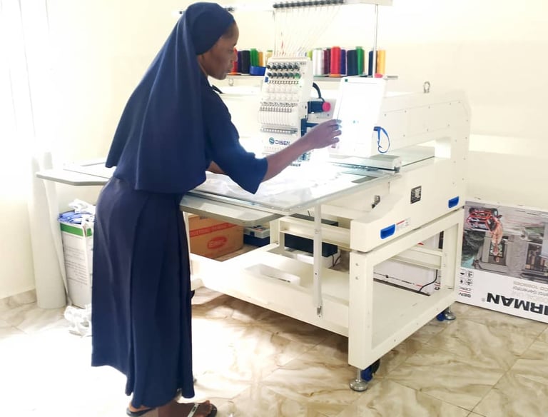 A nun in blue habit operating a white professional multi-needle embroidery machine in a workshop.