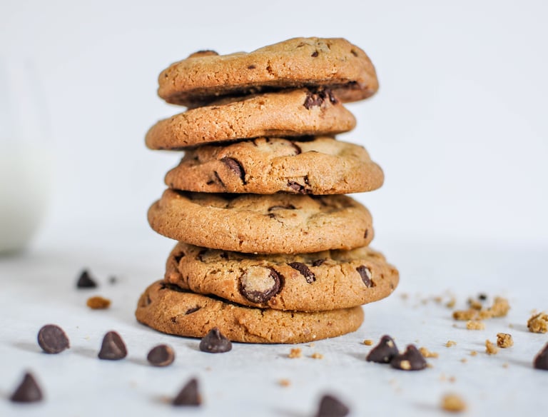 A tall stack of fresh homemade chocolate chip cookies on a white surface with loose morsels.