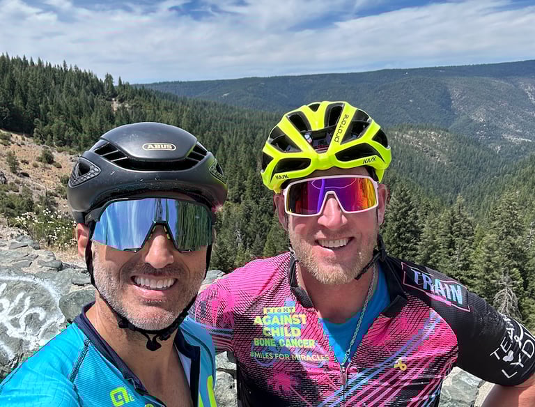 Two cyclists (men) posing for a picture at the top of a mountain range