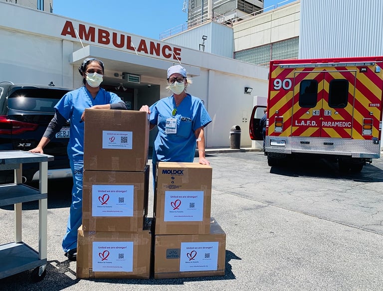 Two healthcare workers in scrubs and masks standing beside stacked supply boxes labeled with heart l
