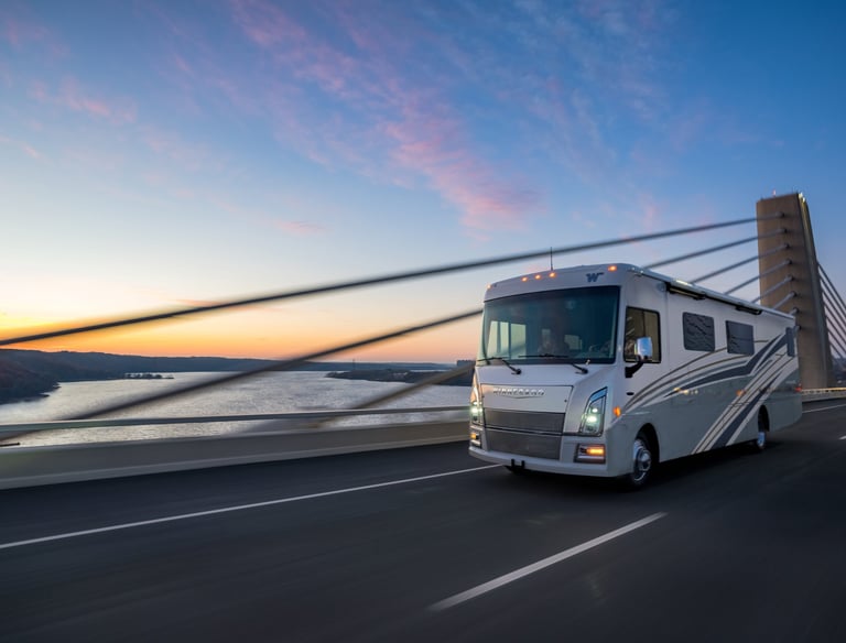A Winnebago Class A motorhome traveling across a suspension bridge at sunset.