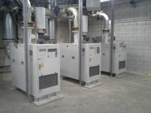 Three industrial air compressor units with ventilation piping in a mechanical room.
