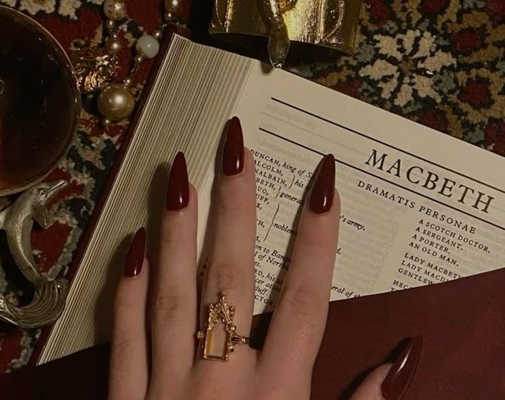 Hand with dark red stiletto nails and gold statement ring resting on Macbeth book.