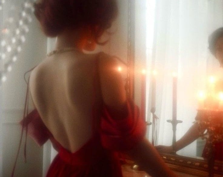 Woman in backless red satin gown facing candlelit mirror, pearl necklace.
