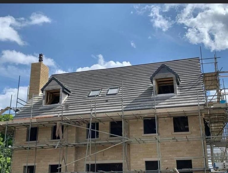 New house construction with a gray tiled roof, stone walls, and scaffolding under a blue sky.
