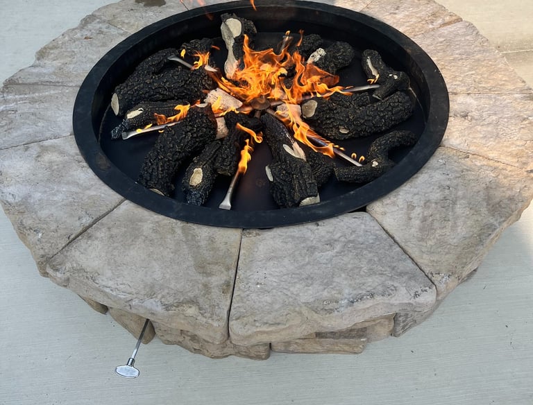 Round outdoor stone gas fire pit with burning ceramic logs on a concrete patio.