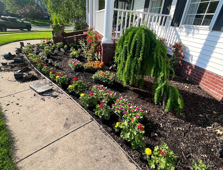 A neatly landscaped garden bed with colorful flowers and mulch along a white house porch and sidewalk.