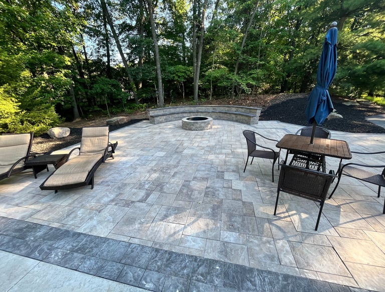 Custom stone paver patio featuring a round fire pit, curved stone bench, and outdoor dining furniture.