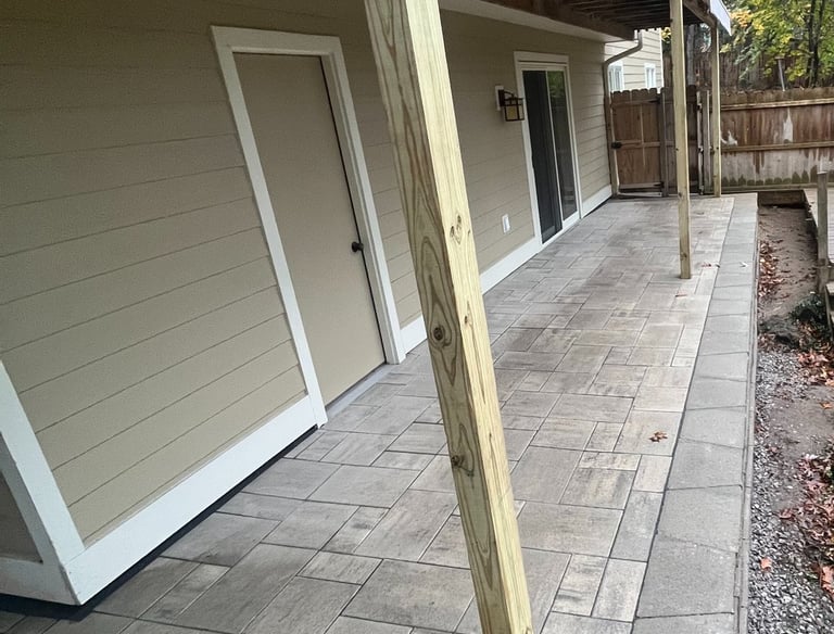 Custom stone paver patio installation under a wooden deck with tan siding and sliding glass doors.