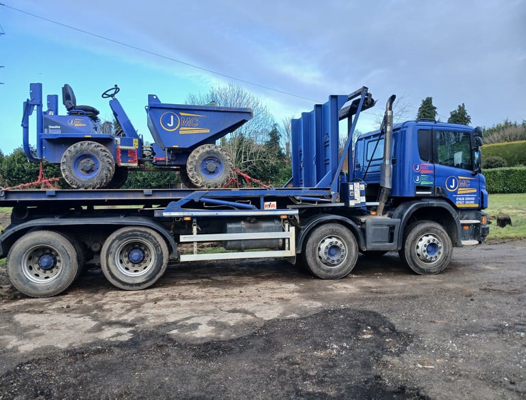 A blue flatbed transport truck carrying a Thwaites site dumper for construction equipment delivery.