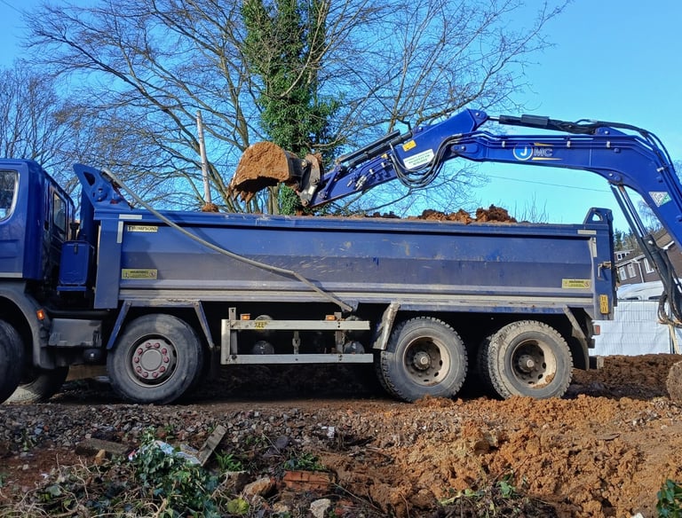A blue grab lorry being loaded with soil by an excavator at a construction site.