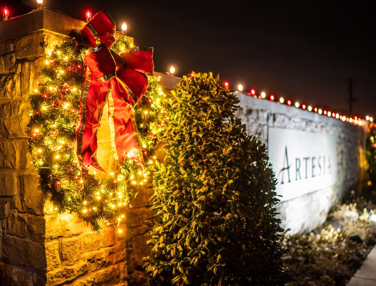 A lit Christmas wreath with a red bow hangs on a stone wall next to sign.