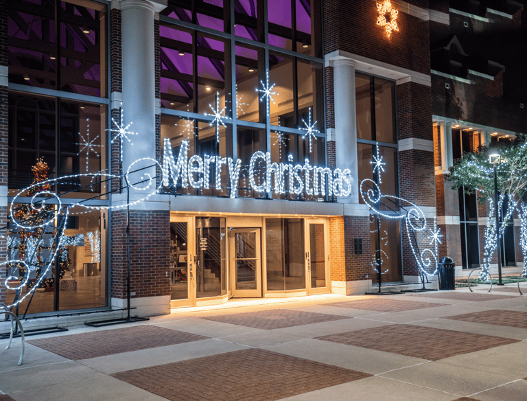Merry Christmas neon sign and white holiday lights decorating a brick building entrance at night.