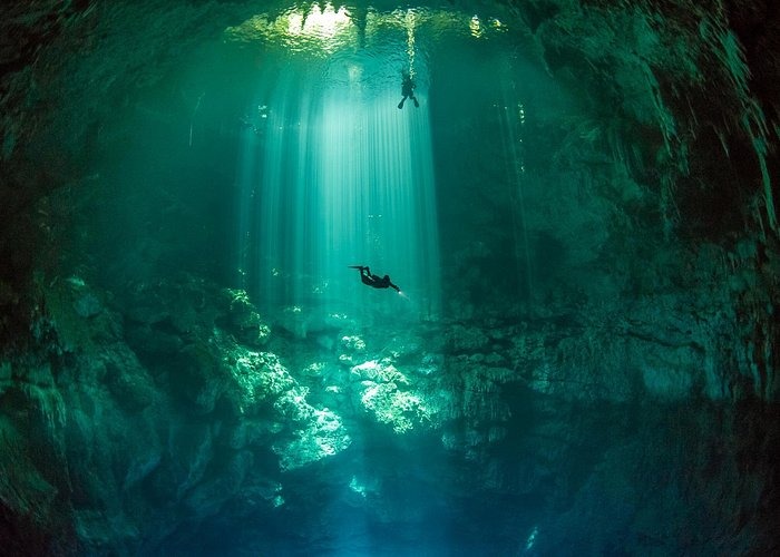 Scuba divers swimming through sunbeams in a deep underwater cenote cave with turquoise water.