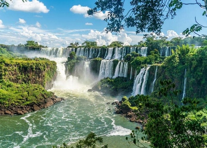 Panoramic view of Iguazu Falls with cascading waterfalls and lush green tropical rainforest under a blue sky.