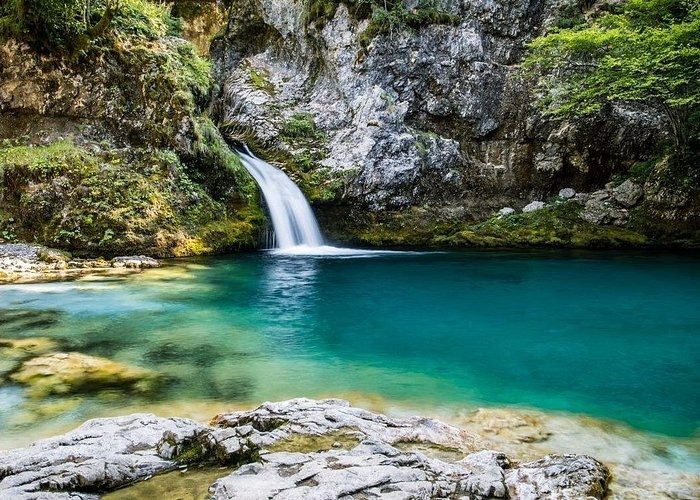 A scenic waterfall flowing into a crystal clear turquoise blue pool surrounded by rocky cliffs and lush green foliage.