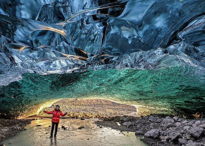 A hiker in a red jacket stands inside a glowing blue ice cave in Iceland with glacial formations.