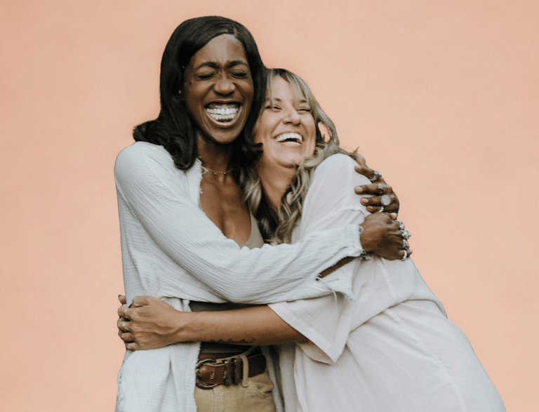 Twee vrouwen lachen en omhelzen elkaar hartelijk tegen een zachte perzikkleurige achtergrond. Ze stralen warmte, verbondenhei