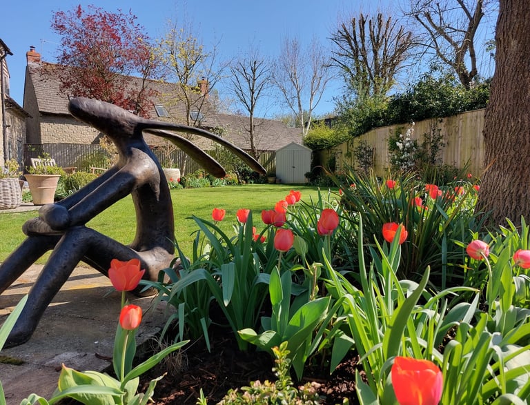 Bronze hare sculpture in a sunny garden with blooming orange tulips and a green lawn.