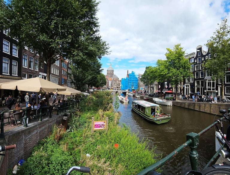 Panoramic view of an Amsterdam canal with a green tour boat, historic canal houses, and outdoor cafes.